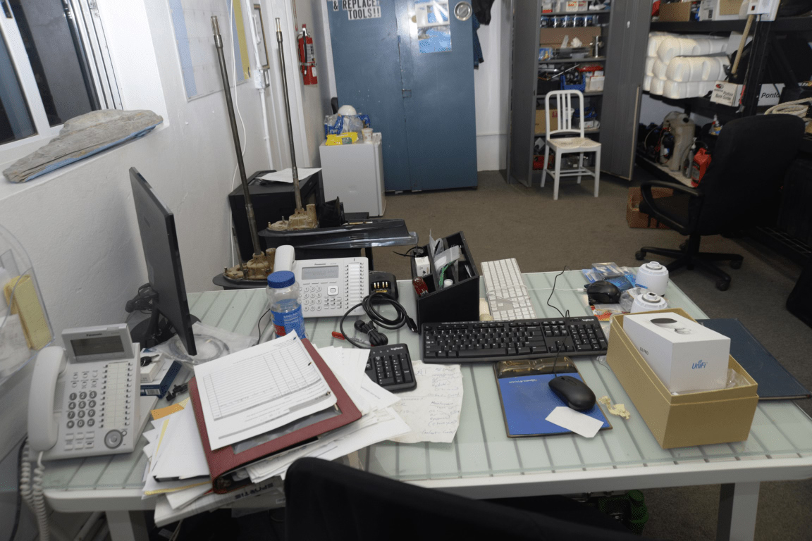 Schreibtisch mit Computermonitor, Tastatur, Maus, Telefonen und Papierstapeln; im Hintergrund Regale mit Kartons und eine blaue Tür.
