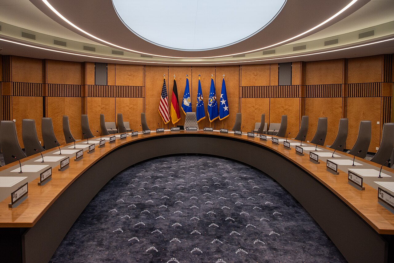 Konferenzraum mit hufeisenförmigem Tisch, Stühlen, Mikrofonen und Flaggen.