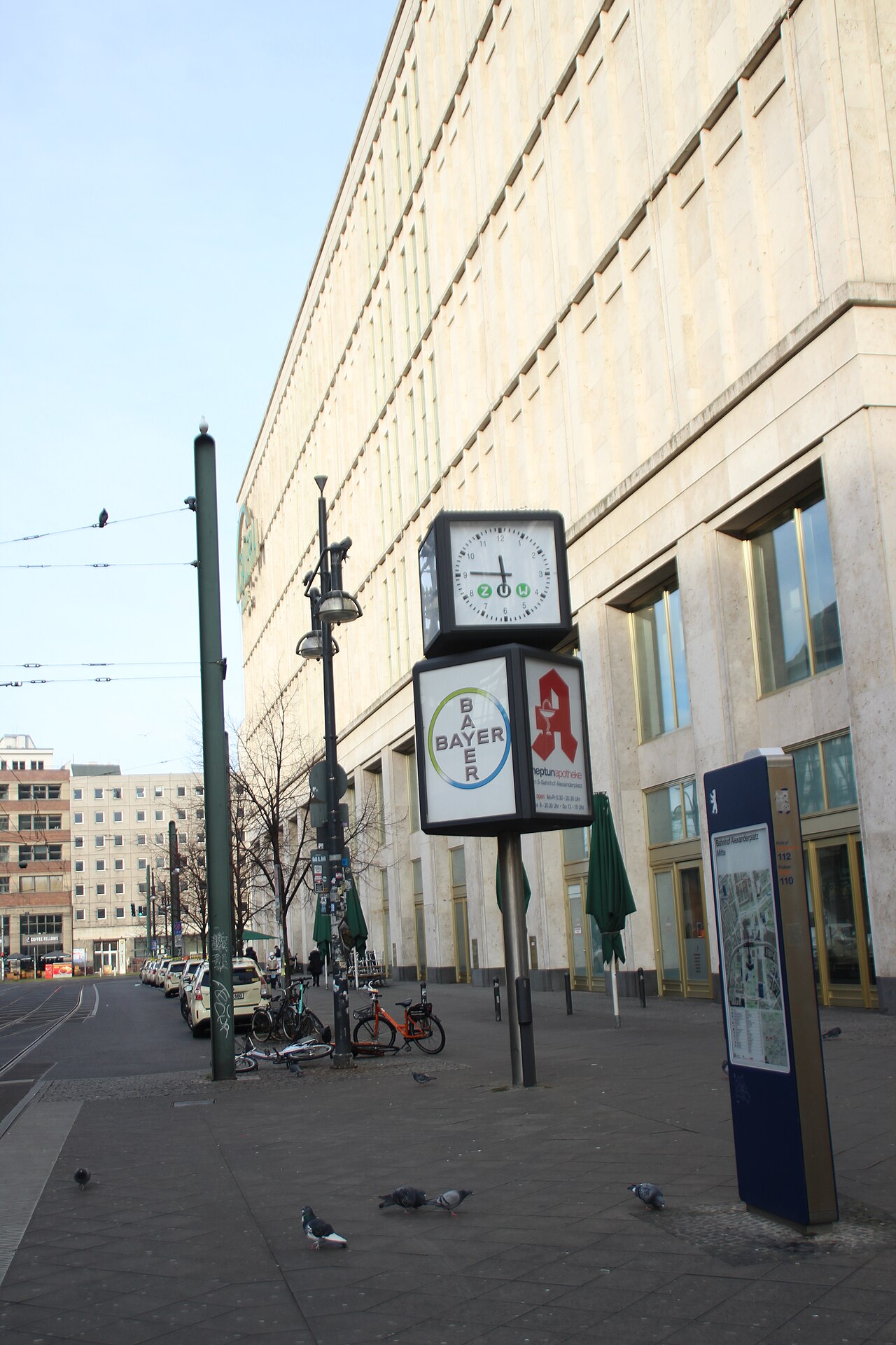 Straßenansicht in Berlin: Uhr mit Werbung für BAYER und neptunapotheke, dahinter Galeria Kaufhof, Straßenbahnschienen und parkende Autos.