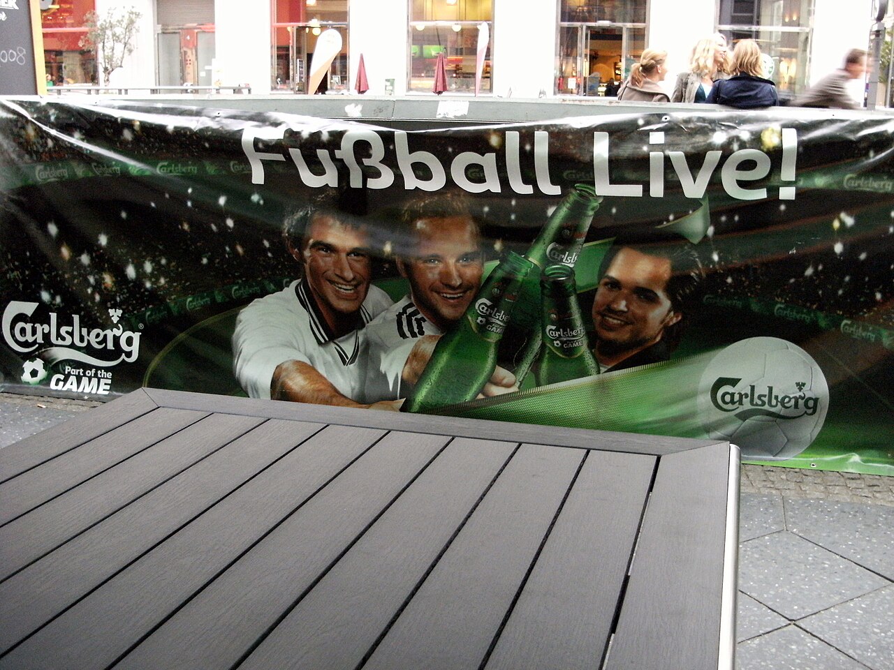Große Werbeplane mit Aufschrift "Fußball Live!" und Carlsberg-Logo, gespannt vor einem Gebäude, im Hintergrund Passanten.