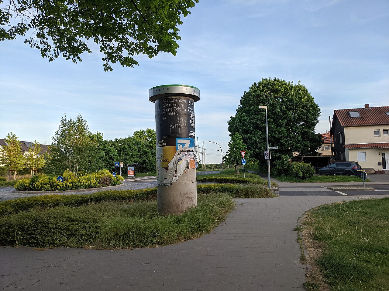 Werbesäule mit Plakaten an einem Gehweg, umgeben von Grün