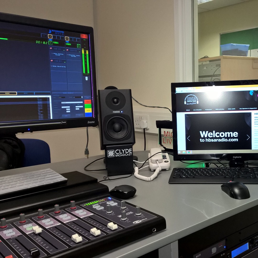 Radiostudio-Arbeitsplatz mit zwei Monitoren, Audio-Schnittsoftware, Mischpult und Lautsprecher